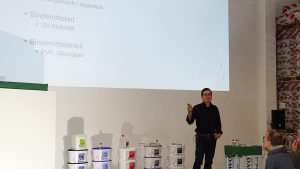 Michael Knapheide steht vor einer weißen Wand mit Beamerbild. Darunter sind diverse Fassadenfarbeimer und Kanister aufgebaut