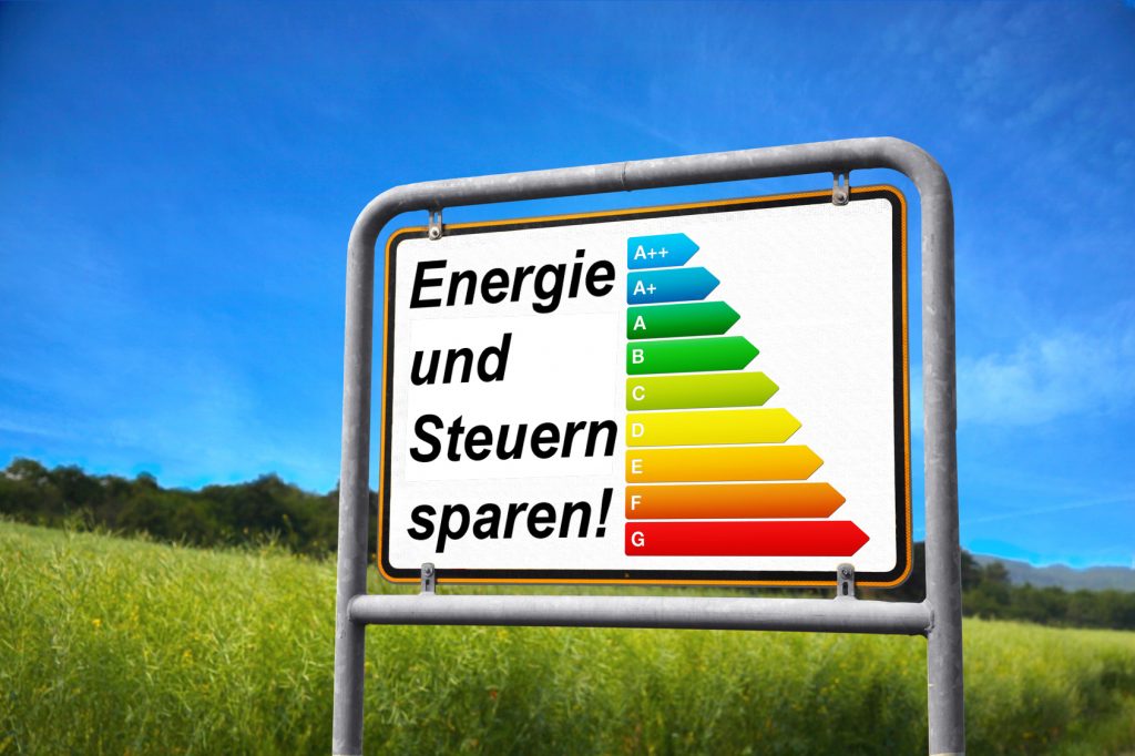 Ein Ortschild, auf dem aber eine Energieampel mit dem Text -Energie und Steuern sparen!- zu sehen ist - im Hintergrund Wiese, Bäume und Himmel