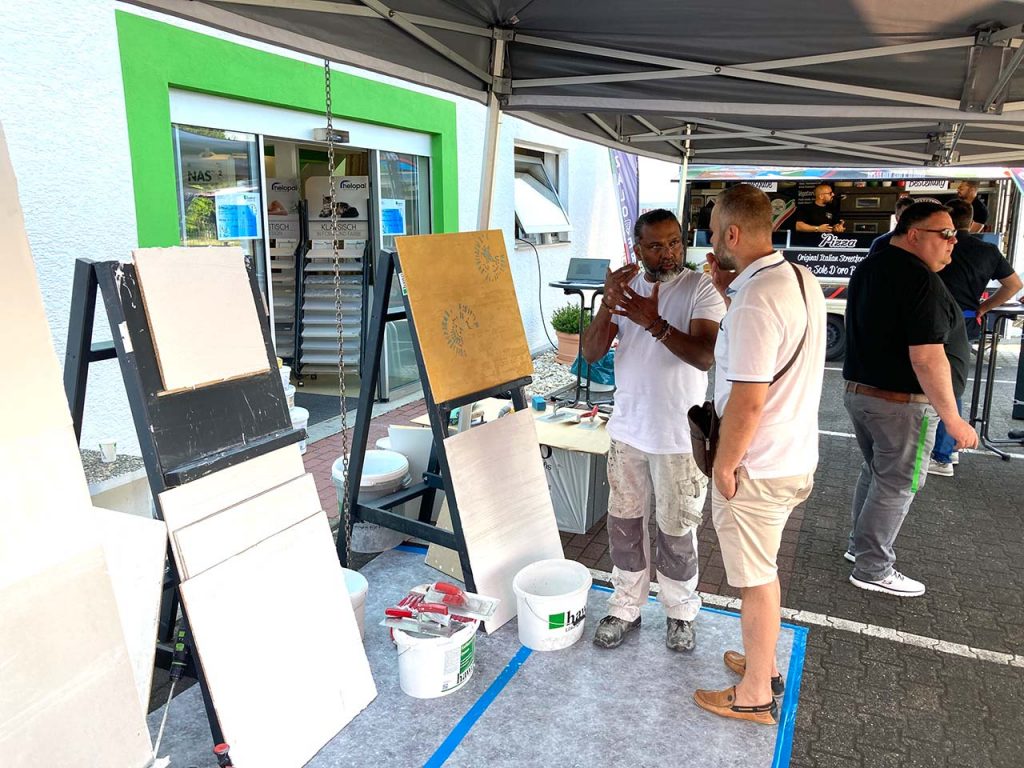 Kreativecke im Außenbereicht - Zwei Staffeleien mit Musterplatten - Dilip erklärt einem Kunden etwas