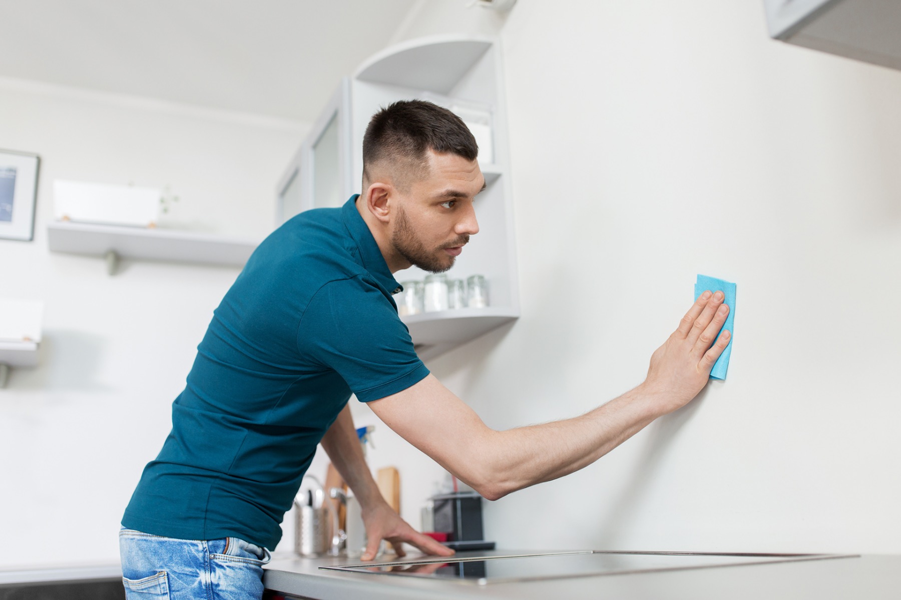 Mann wischt mit einem blauen Lappen an einer weißen Wand oberhalb der Küchen-Arbeitsplatte - hinder dem Herd